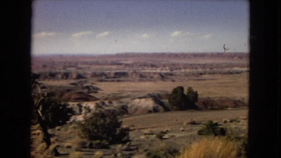1967 Arizona home-movie frame showing dry open land, low rock formations, scrub vegetation, a small tree at left, and distant mesa-like terrain.