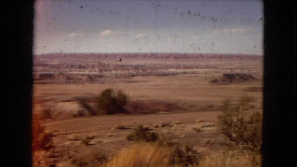 Faded 1967 Arizona 8mm travel frame showing open desert grassland, blurred shrubs, low distant hills, and a broad sky with warm color shift at the frame edge.