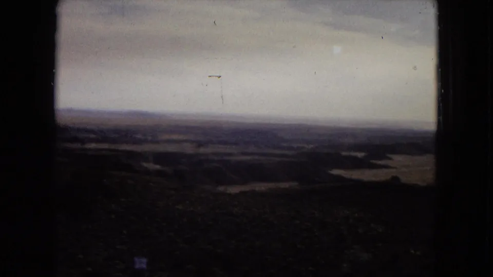 1967 Arizona 8mm frame looking across a dark foreground slope toward a broad arid valley, low hills, and a hazy pale sky.