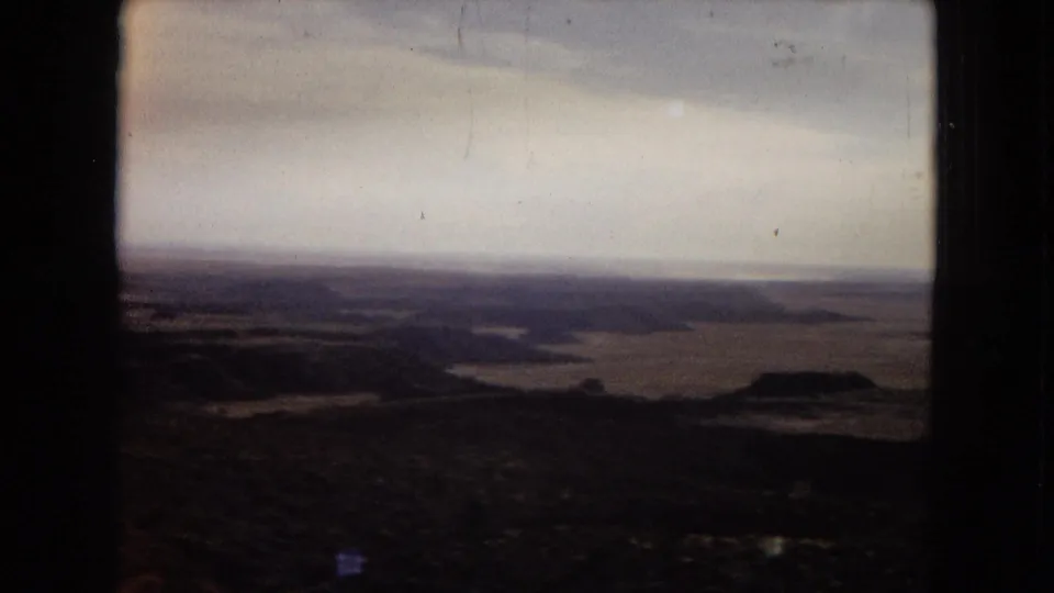 1967 Arizona home-movie frame showing a broad desert valley panorama, dark foreground landforms, distant horizon, and pale clouded sky with visible 8mm wear.