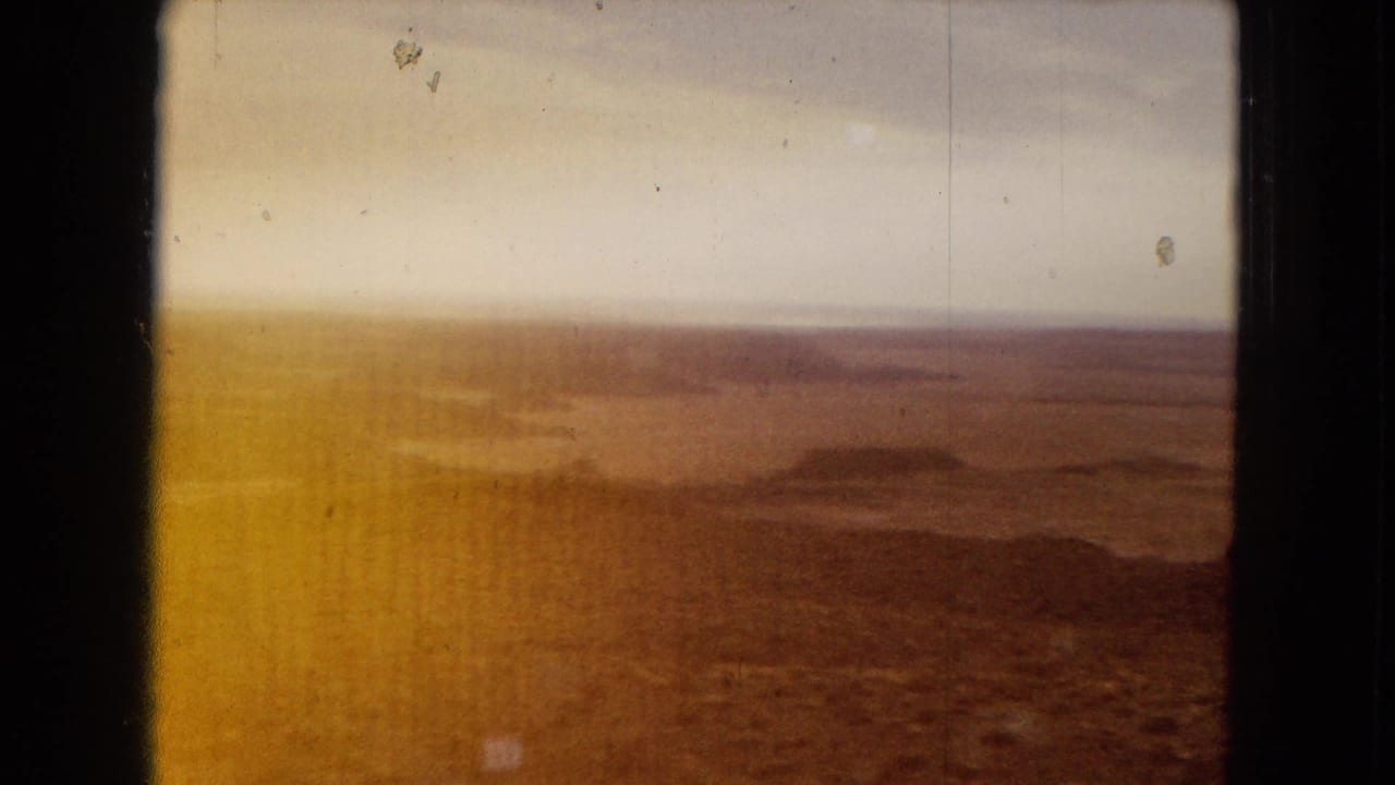 1967 Arizona 8mm frame showing a hazy arid plain with distant low hills, pale sky, warm color fade, and dark border framing the view.