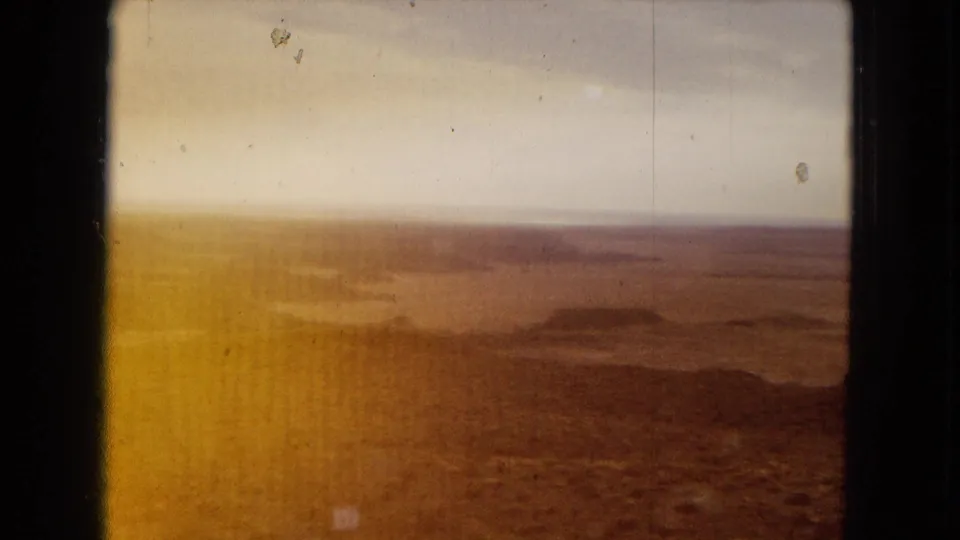 1967 Arizona 8mm frame showing a hazy arid plain with distant low hills, pale sky, warm color fade, and dark border framing the view.