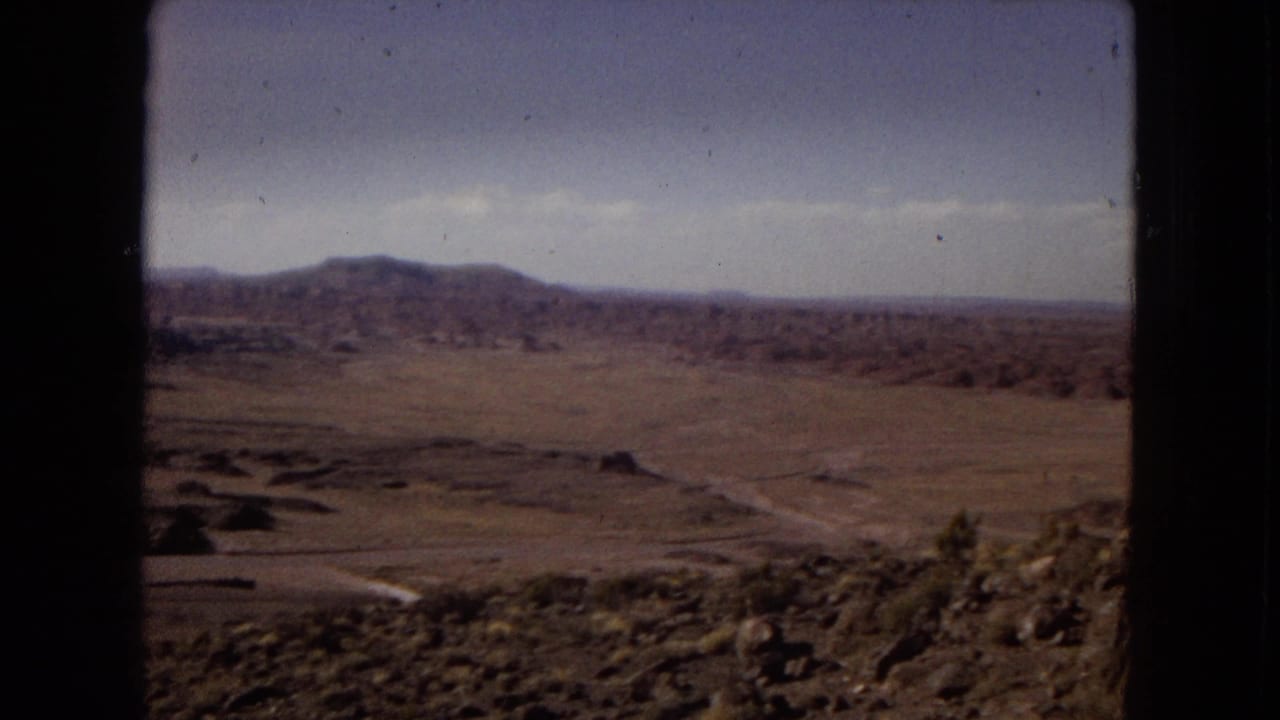 1967 Arizona 8mm view across a dry valley with rocky foreground terrain, scrub, low ridges, distant mountains, and a dark vertical frame edge.