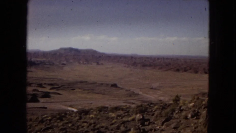 1967 Arizona 8mm view across a dry valley with rocky foreground terrain, scrub, low ridges, distant mountains, and a dark vertical frame edge.