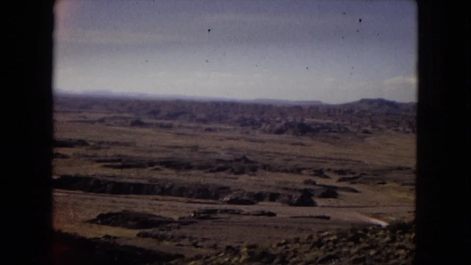 1967 Arizona 8mm frame showing a broad desert valley, scattered scrub, low hills, distant ridgeline, and a pale winding road or wash through the landscape.