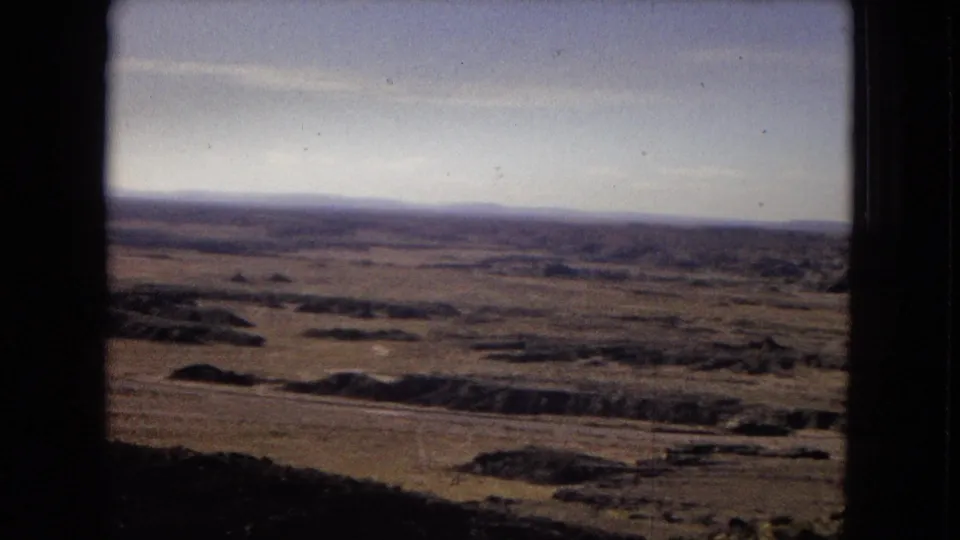 1967 8mm home-movie view of an arid Arizona plain with scattered boulders, distant low mountains, pale blue sky, and dark vehicle-window framing.