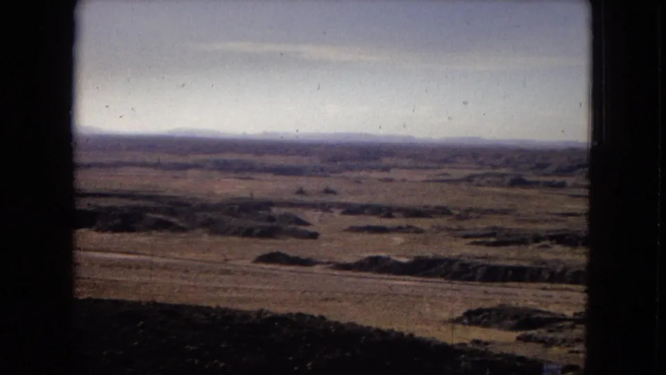 1967 Arizona travel footage showing open desert plains, low rocky rises, and distant ridges under a bright sky, partially bordered by dark vehicle-window edges.