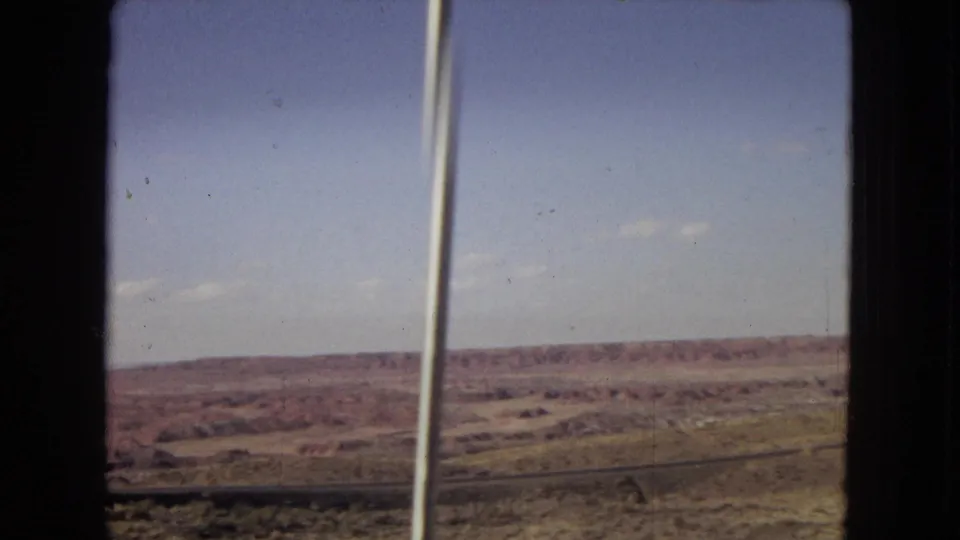 1967 8mm vehicle-shot view across Arizona desert terrain with a vertical pole near the foreground, low mesas in the distance, and a clear blue sky.