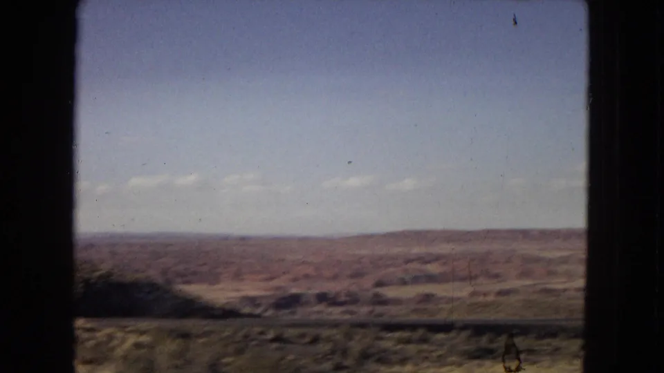 1967 Arizona home-movie frame showing rolling arid ground, distant hills or mesas, scattered shadows, and a pale blue sky from a moving vehicle perspective.