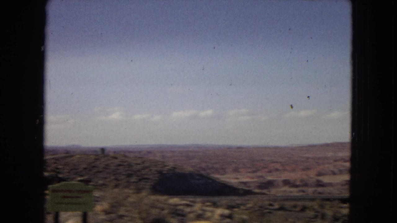 1967 Arizona home-movie view of open desert terrain and distant low hills, framed by the dark edge of a vehicle window with a green roadside sign partly visible.