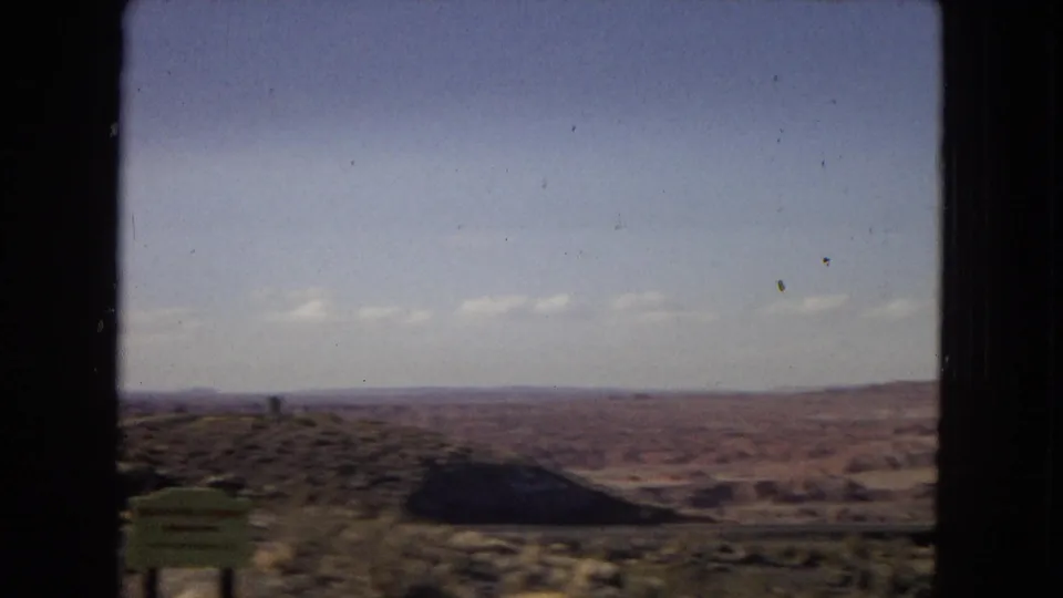 1967 Arizona home-movie view of open desert terrain and distant low hills, framed by the dark edge of a vehicle window with a green roadside sign partly visible.