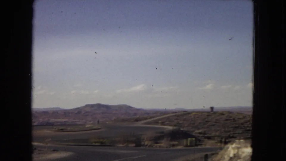1967 Arizona road-view frame showing a paved route curving through arid terrain, with low red-brown hills and a dark vehicle-window border around the scene.