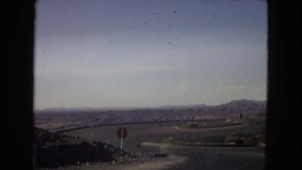 1967 Arizona home-movie frame from inside a vehicle, showing a paved desert road, distant rocky hills, and a red stop sign near the lower left edge.