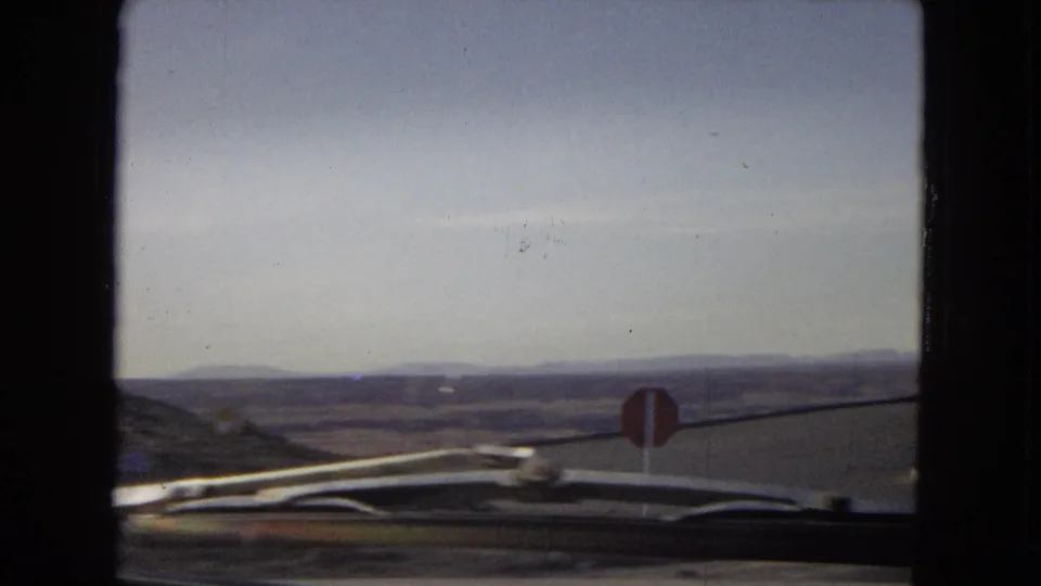 1967 Arizona driving view through a windshield, with wiper blades and dashboard edge visible in front of a paved desert road and a red stop sign.