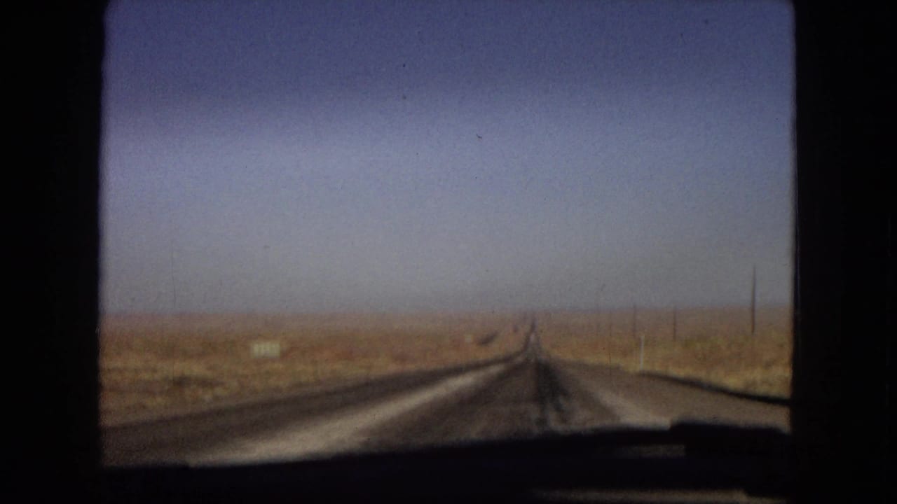 1967 Arizona home-movie frame showing a straight open road receding through dry desert terrain, filmed from inside a moving vehicle with window edges visible.