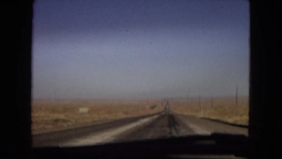 1967 Arizona home-movie frame showing a straight open road receding through dry desert terrain, filmed from inside a moving vehicle with window edges visible.
