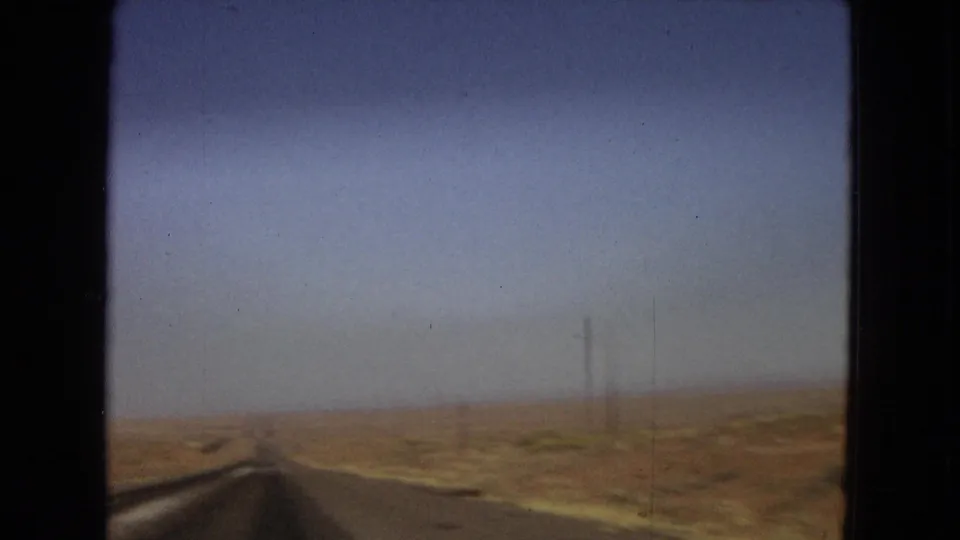 1967 Arizona home-movie frame looking past the vehicle edge toward a paved desert road, dry grassland, utility poles, and a hazy horizon.