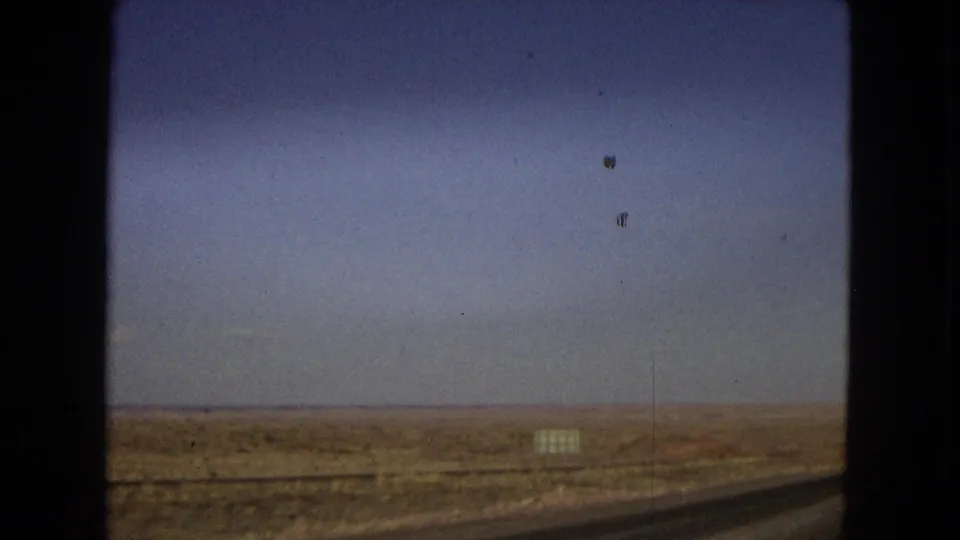 1967 Arizona 8mm frame showing the desert roadside from inside a moving vehicle, with a broad blue sky, low arid horizon, roadside sign, and part of the road at lower right.