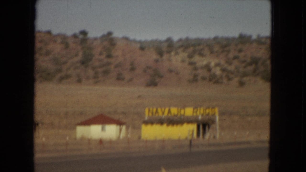1967 Arizona home-movie frame showing a yellow roadside stand with a "NAVAJO RUGS" sign, a small white building with a red roof, and dry hills behind them.