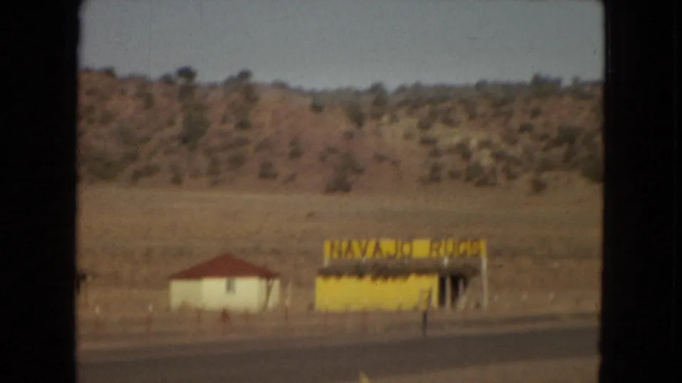 1967 Arizona home-movie frame showing a yellow roadside stand with a "NAVAJO RUGS" sign, a small white building with a red roof, and dry hills behind them.