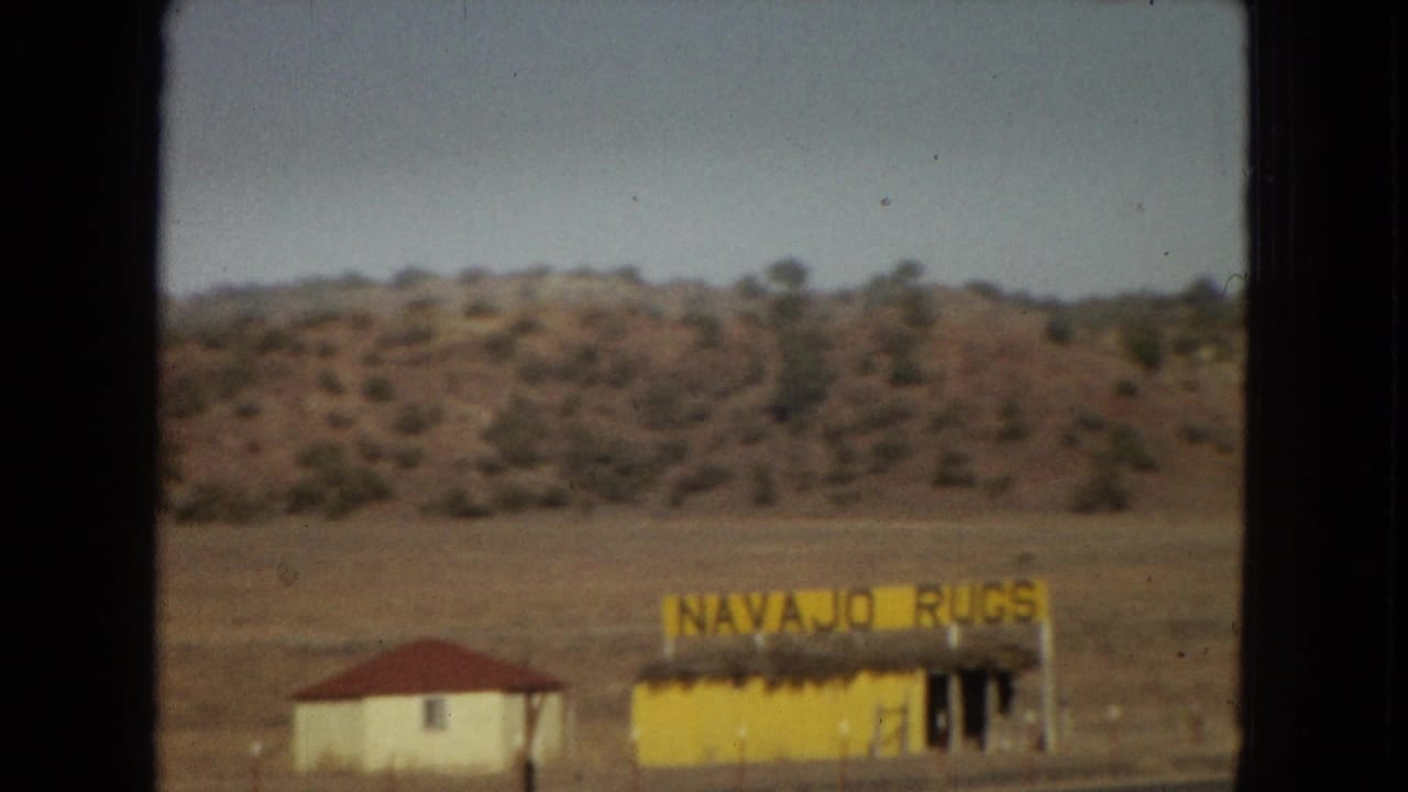 1967 Arizona road-trip frame with the yellow "NAVAJO RUGS" roadside building, a neighboring white structure, dry open ground, and low desert hills.
