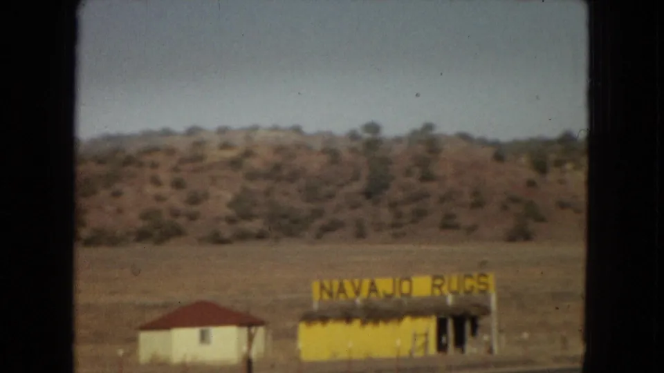 1967 Arizona road-trip frame with the yellow "NAVAJO RUGS" roadside building, a neighboring white structure, dry open ground, and low desert hills.
