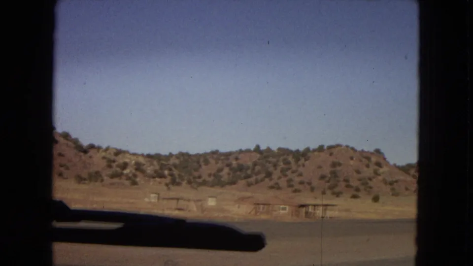 1967 Arizona home-movie frame looking from inside a vehicle toward dry hills, sparse roadside structures, and a dark windshield or dashboard edge.
