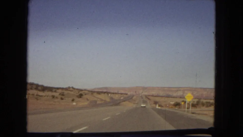 1967 Arizona road-trip frame from inside a moving car showing an open paved highway, distant vehicles, roadside signs, and dry desert hills.