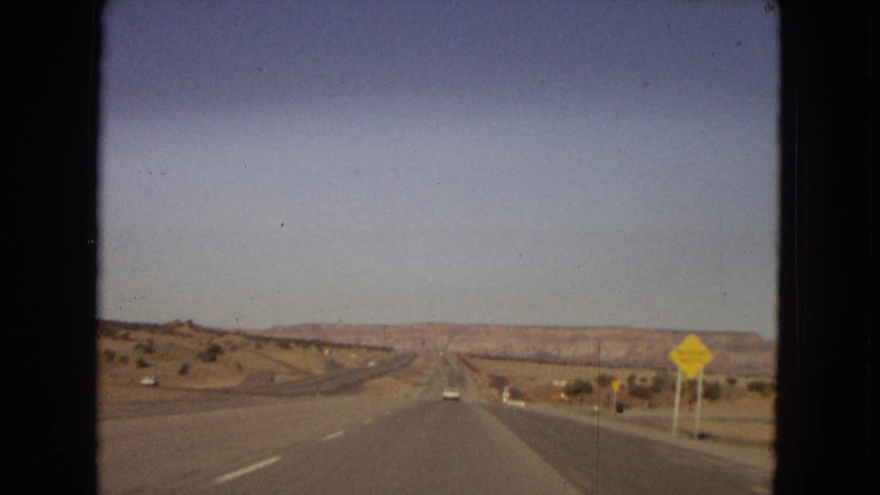 1967 home-movie view from a moving car on a straight desert highway in the United States, with distant mesas or low mountains and a yellow traffic warning sign at the roadside.