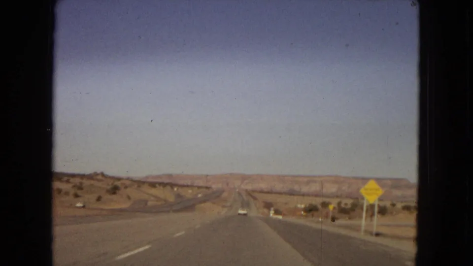 1967 home-movie view from a moving car on a straight desert highway in the United States, with distant mesas or low mountains and a yellow traffic warning sign at the roadside.