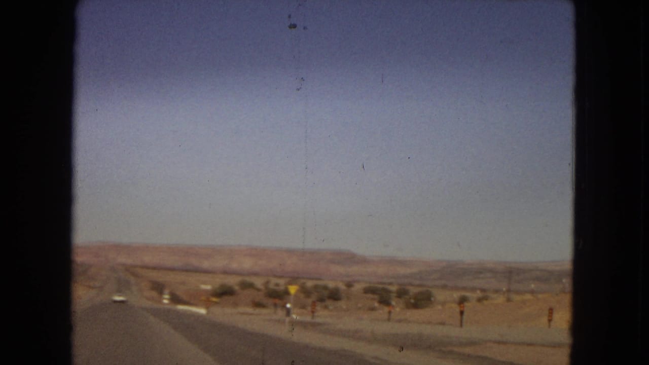 Moving-car view across an arid western highway landscape, with a roadside merge or warning sign, sparse desert vegetation, and low mesas under a clear sky.