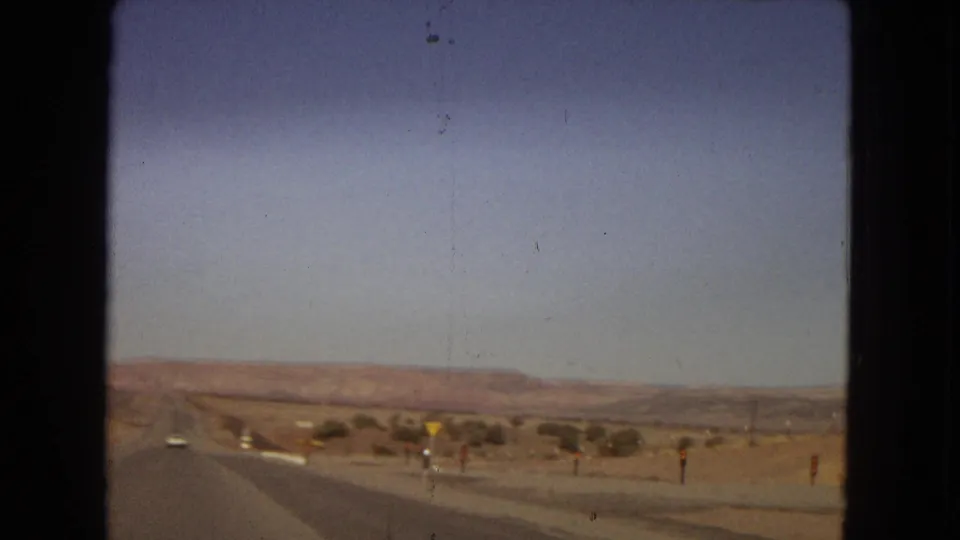 Moving-car view across an arid western highway landscape, with a roadside merge or warning sign, sparse desert vegetation, and low mesas under a clear sky.