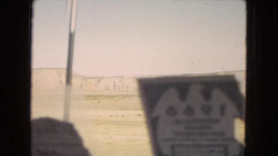 Soft, partially obstructed 8mm frame from inside a moving car, showing a blurred foreground object, windshield edge, pale desert cliffs, and bright sky beyond.