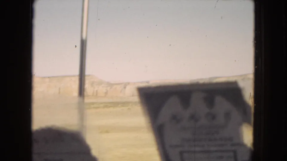 1967 home-movie frame from a moving vehicle in the United States, showing pale desert terrain, distant mesas, and a blurred interior card or sign near the window.