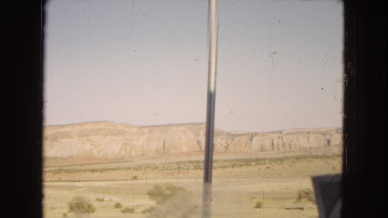 1967 moving-vehicle view across a dry desert landscape, with layered mesas in the distance and vertical vehicle-window framing in the foreground.