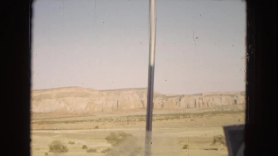 1967 moving-vehicle view across a dry desert landscape, with layered mesas in the distance and vertical vehicle-window framing in the foreground.