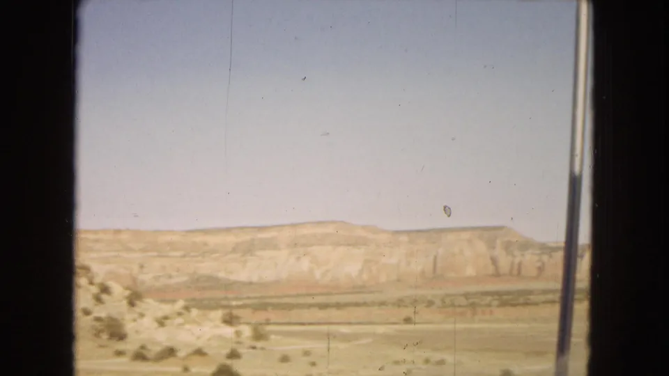 1967 home-movie view from a moving vehicle, looking across scrubby desert ground toward a pale plateau under a clear daytime sky.