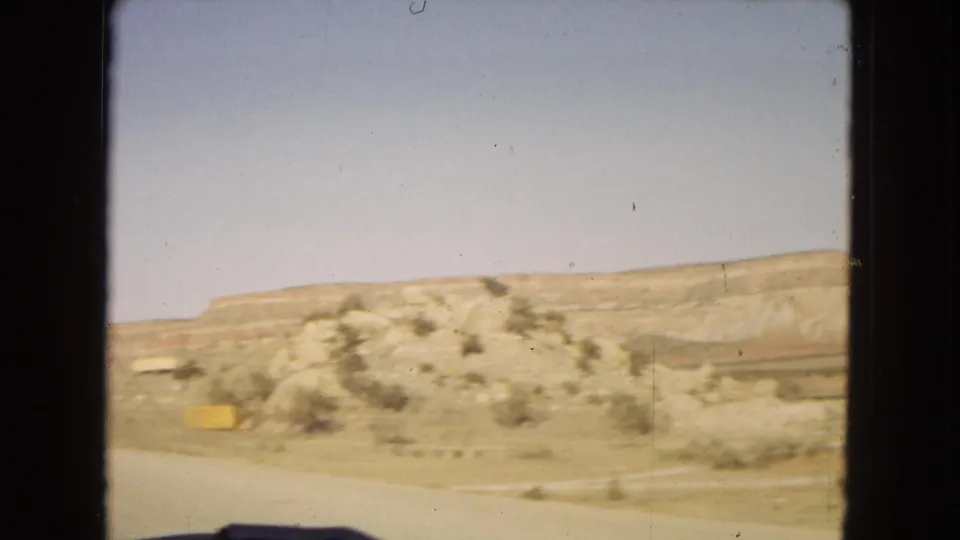 1967 moving-car frame showing a low rocky desert hill, dry roadside ground, sparse brush, and pale mesa formations in the background.