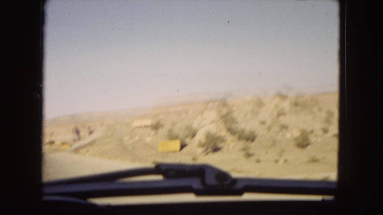 1967 Arizona road-trip footage appears to show arid roadside hills and a yellow sign viewed through a moving car window, with the windshield wiper and dark film gate visible.