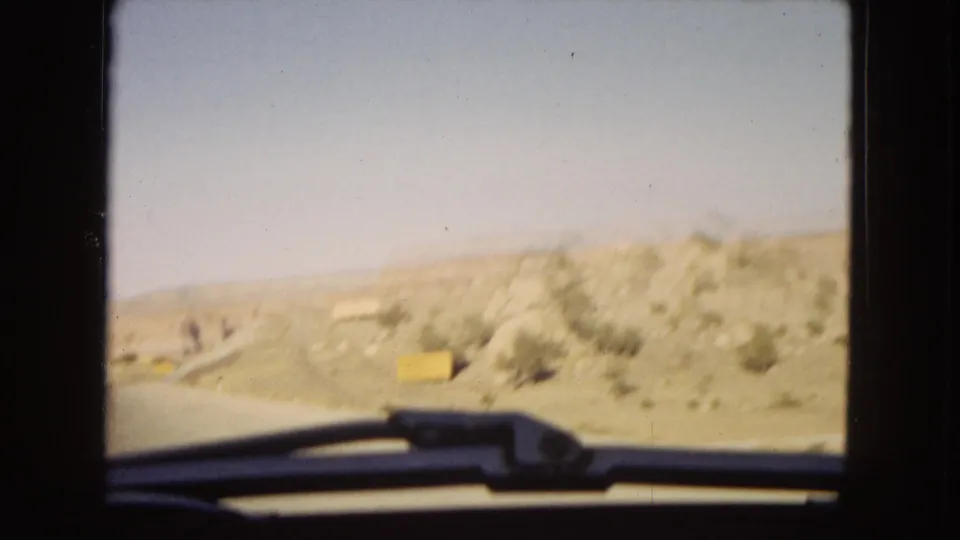 1967 Arizona road-trip footage appears to show arid roadside hills and a yellow sign viewed through a moving car window, with the windshield wiper and dark film gate visible.