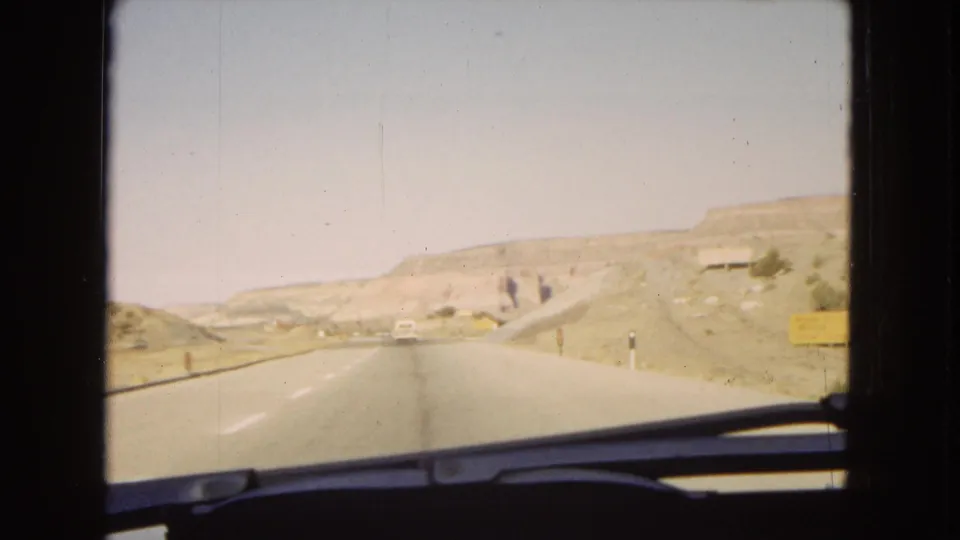 1967 Arizona footage shows a paved desert highway stretching through low dry hills, with distant roadside figures, signs, and the car windshield edge visible in the foreground.