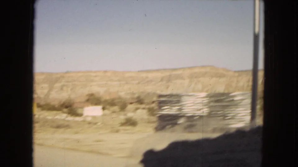 A moving-car view in the 1967 Arizona road-trip clip shows blurred scrubland, low desert hills, a roadside sign, and a vertical pole passing close to the frame.