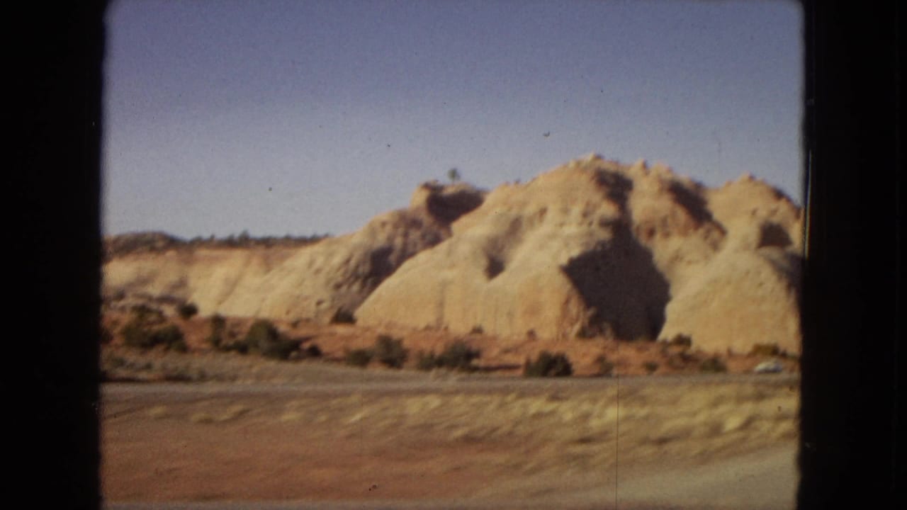 1967 8mm home-movie view from a moving car showing tan sandstone formations and sparse desert roadside vegetation in Arizona.