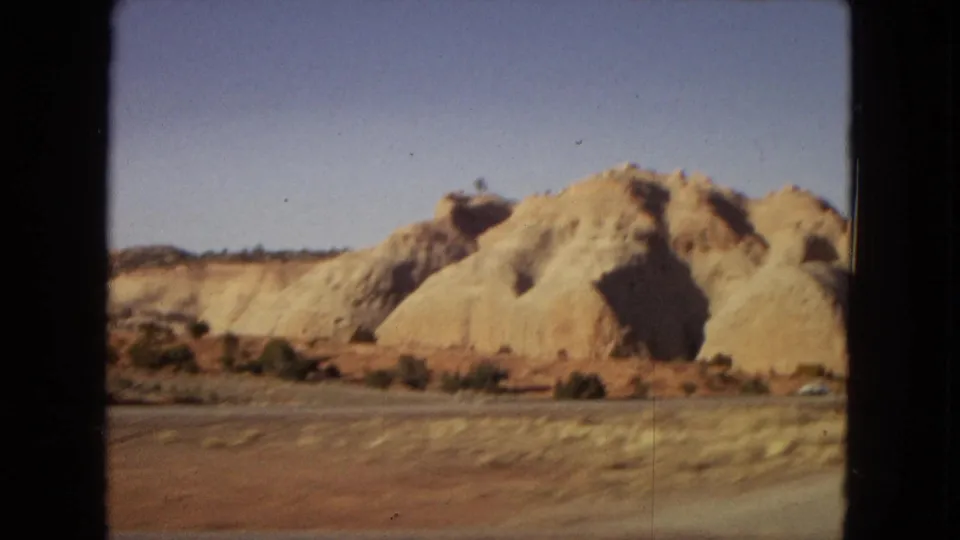 1967 8mm home-movie view from a moving car showing tan sandstone formations and sparse desert roadside vegetation in Arizona.
