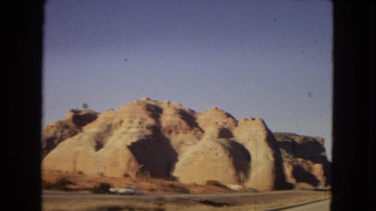 Arizona desert highway footage from 1967 showing rounded tan rock formations, sparse brush, and a small white car near the roadside.