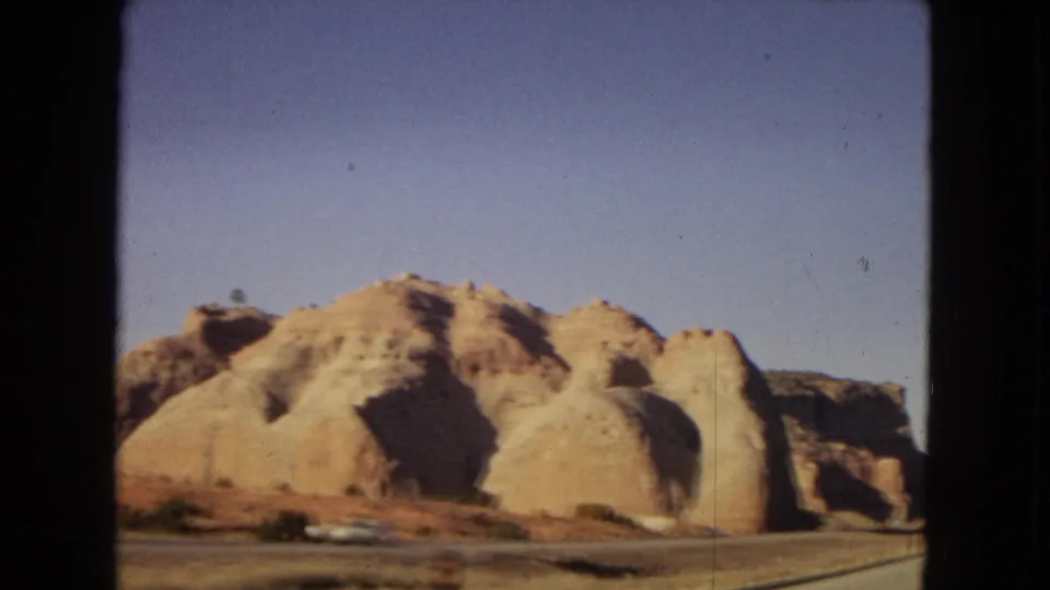 Arizona desert highway footage from 1967 showing rounded tan rock formations, sparse brush, and a small white car near the roadside.