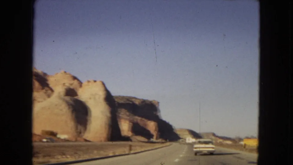 Moving-vehicle view on an Arizona desert highway in 1967, with a white car ahead and tan rock formations along the roadside.