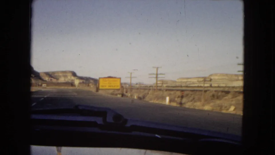1967 8mm road view through a car windshield showing a desert highway, utility poles, distant mesas, and a yellow road sign in Arizona.
