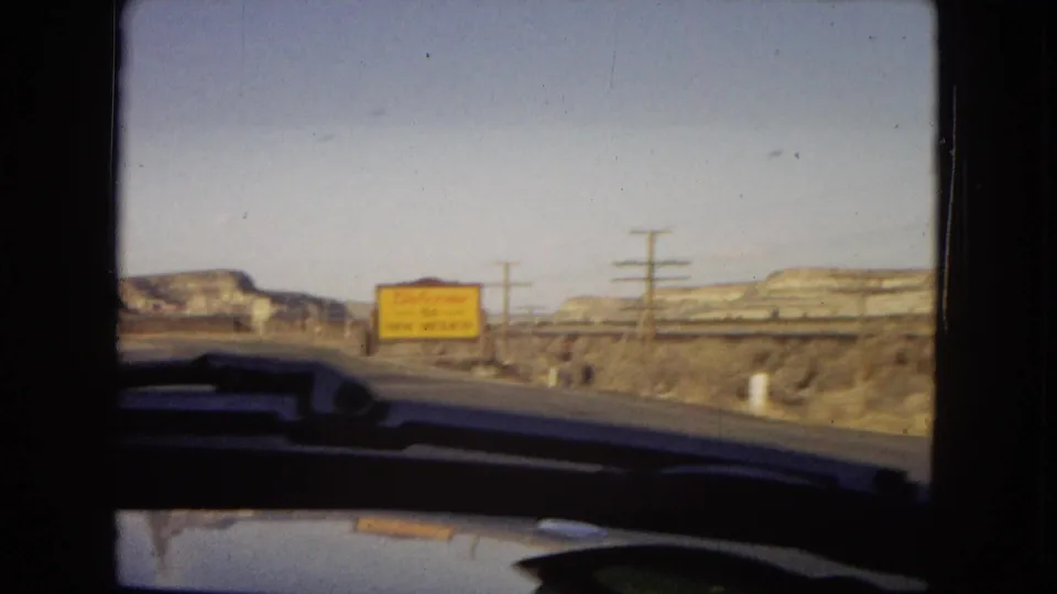 1967 8mm road-trip view from inside a moving car approaching a yellow Welcome to New Mexico roadside sign with mesas and utility poles beyond.