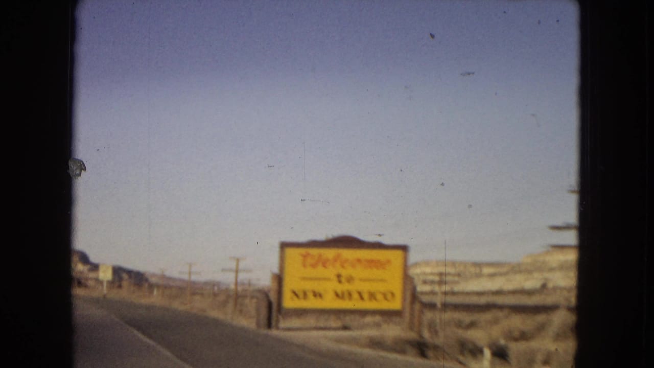 A yellow Welcome to New Mexico sign stands beside the highway as the moving-car view continues through an open Southwestern desert landscape.